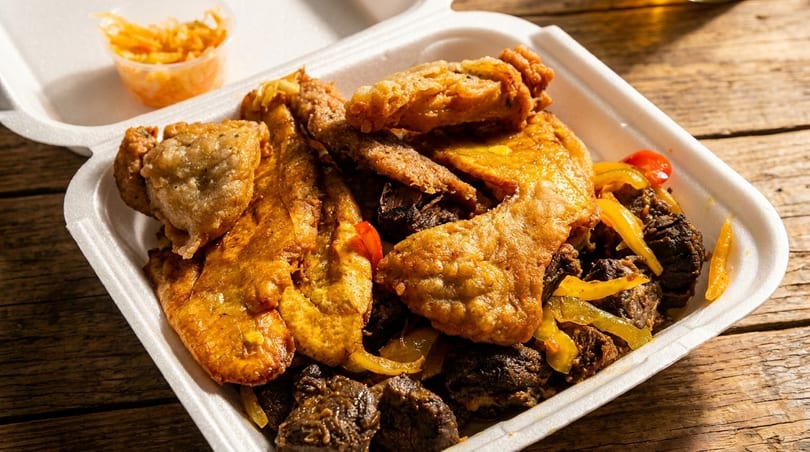 Fried plantains and meat with peppers in a white takeout container on a wooden table.