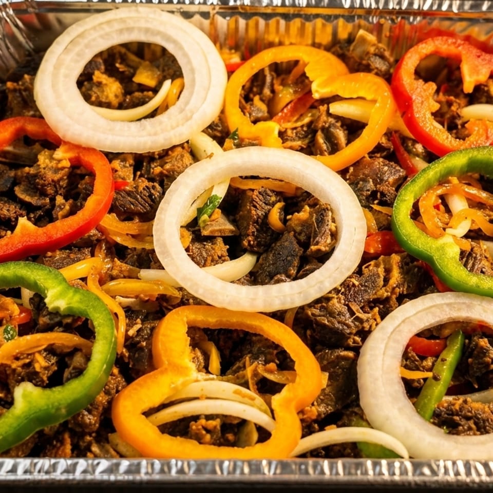 A foil tray filled with diced meat, topped with colorful bell pepper rings and white onion slices.