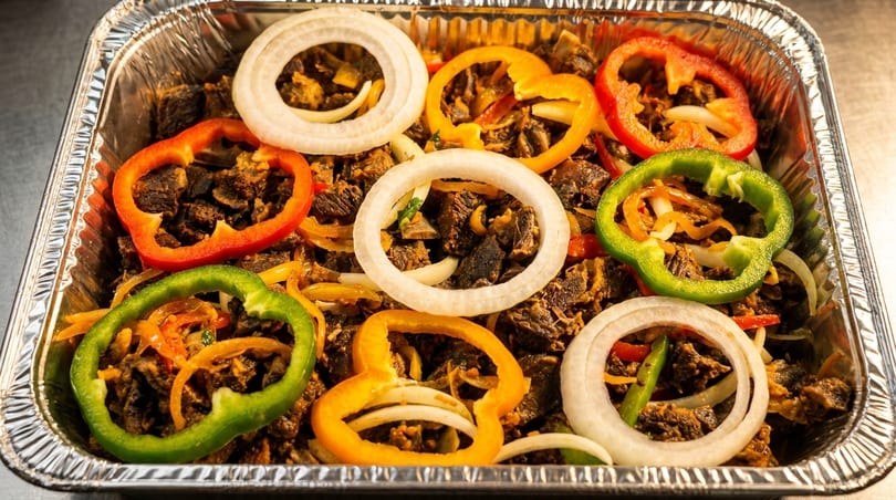 A foil tray filled with diced meat, topped with colorful bell pepper rings and white onion slices.